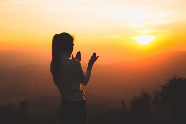Silhouette of a person standing with hands up, facing a vibrant sunset over a mountain landscape.