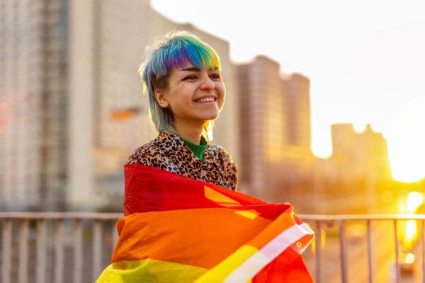Person smiling, wrapped in a rainbow flag, with a colorful hairstyle and a cityscape in the background.