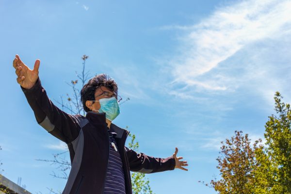 Person with outstretched arms wearing a mask, outdoors under a blue sky with trees.