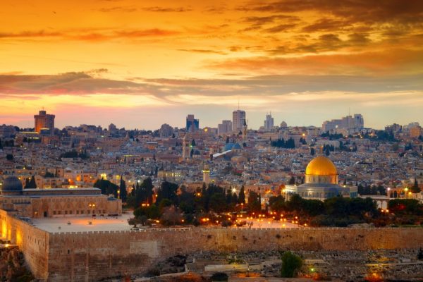 A panoramic view of Jerusalems skyline at sunset, featuring the Dome of the Rock with a vibrant orange sky.