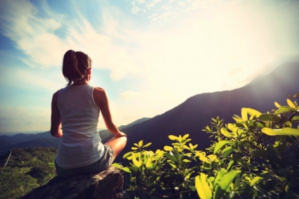 A woman sits on a rock, meditating while facing a scenic mountain view under a bright sky.