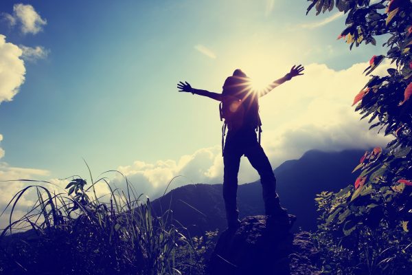 Silhouette of a person with arms outstretched on a mountain, sun shining through, surrounded by lush greenery.