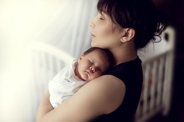 Mother holding a sleeping baby, standing peacefully by a crib with soft, natural lighting.