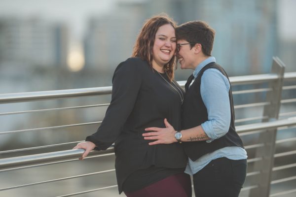 Two people share a playful moment on a bridge, smiling and standing close, with a cityscape in the background.