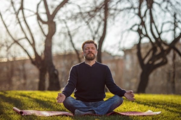 Man meditating in a park, sitting cross-legged on a mat with eyes closed, surrounded by trees.