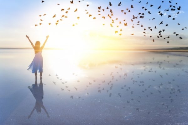 A person stands with arms raised on a reflective beach at sunset, surrounded by a flock of birds in flight.
