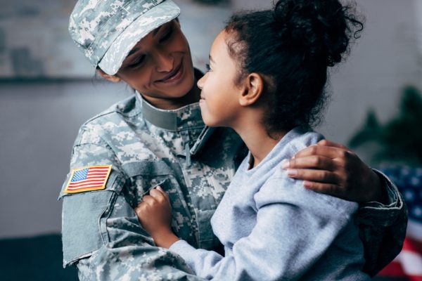 A soldier in uniform embraces a young girl, both smiling warmly at each other.