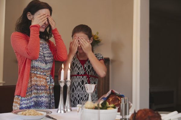 Two people standing with closed eyes, covering their faces, in front of a table set with candles and food.