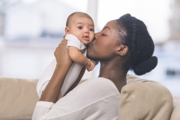 A woman kisses a baby on the cheek while holding them in her arms, sitting on a couch indoors.