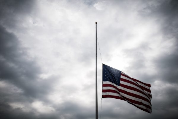 US flag at half-mast against a cloudy sky, symbolizing mourning or respect.