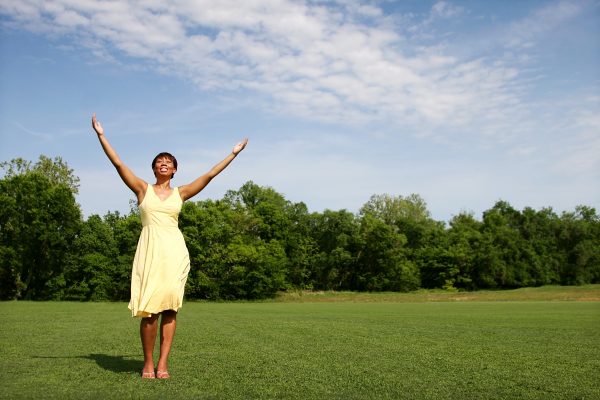 Woman in yellow dress standing with arms raised on a grassy field, surrounded by trees under a partly cloudy sky.