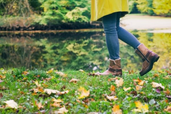 Person in jeans and brown boots walking near a lake, surrounded by autumn leaves and trees.