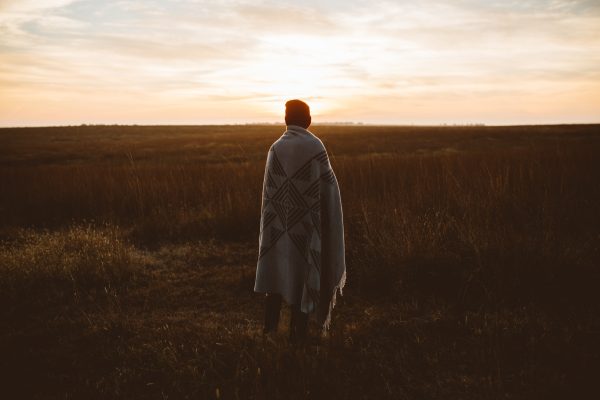 Person wrapped in a blanket, standing in a field, gazing at a sunrise under a cloudy sky.
