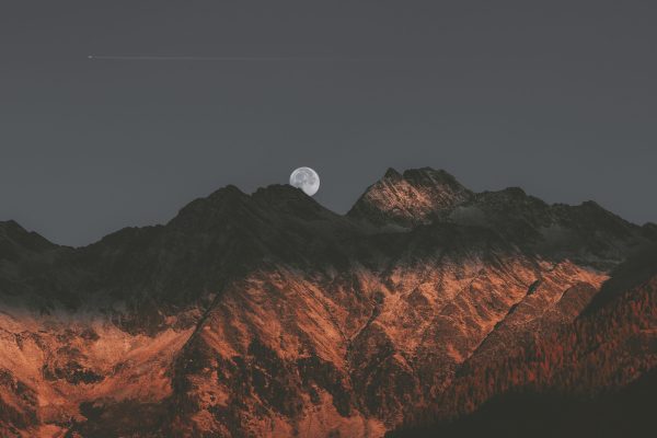 Full moon rising over dark mountain peaks with a red-orange glow and a jet streaking across the night sky.