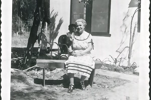 Elderly woman in a dress sits on a bench outside, with a young child behind her near a house.