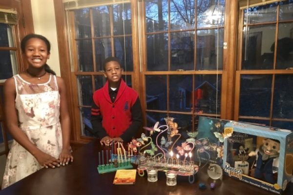 Two children smiling by a table with a menorah and Hanukkah gifts near a window at dusk.