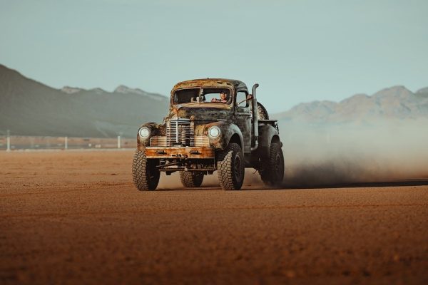 An old rusty pickup truck drives fast across a dusty desert with mountains in the background.