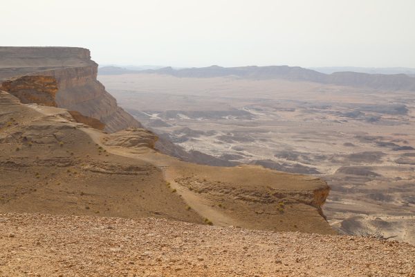 Vast desert landscape with rocky cliffs and distant hazy horizon under a pale sky.