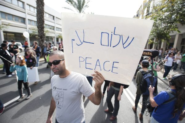 A man at a street protest holds a sign reading PEACE in English and Hebrew.