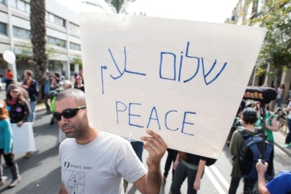 Person holding a sign with Peace written in English and Hebrew at a street rally with people in the background.
