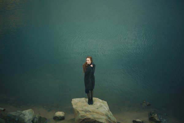 A woman in a black coat stands on a rock by a calm, blue body of water, surrounded by a few scattered stones.