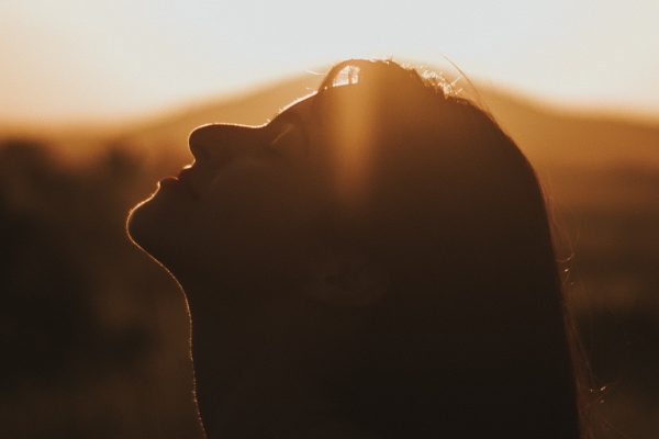 Silhouette of a person facing the sun with closed eyes, in a serene outdoor setting with a warm, glowing sky.