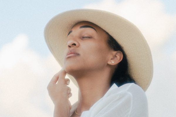 light brown skinned person with eyes closed and white shirt on and beige sun hat with head slightly lifted looking peaceful before light blue sky and clouds