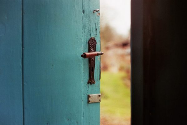 A slightly open blue door with an antique handle, revealing a blurred view of a garden outside.
