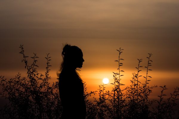 woman in silhouette standing straight looking ahead with setting sun and dark gray sky in background and some trees