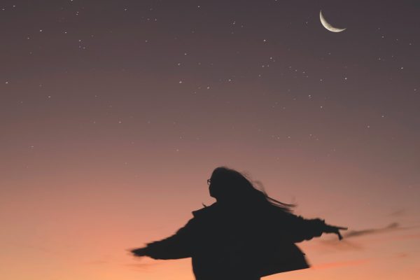 woman in silhouette with arms outspread and hair blowing standing or spinning or dancing in front a purple pink night sky with silver of new moon and stars