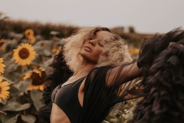 black woman with frizzy blonde hair and wing-like black cape with arms spread in field of sunflowers eyes closed