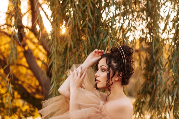 light brown skinned woman with curly dark hair dressed in gauzy gown looking contemplative in halo-like crown with hair pinned up and curls coming down in field with tree and sunlight