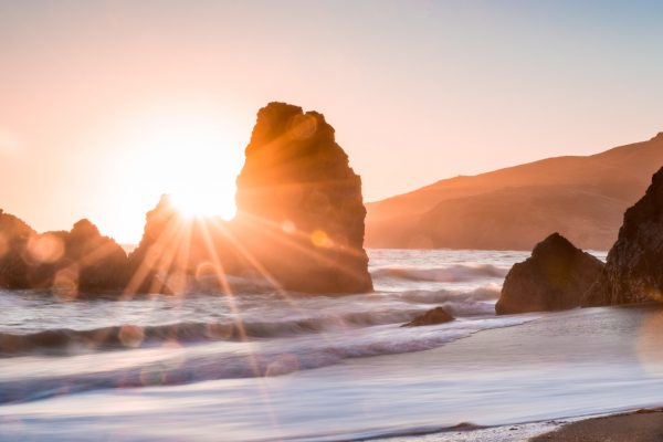 Sunset over ocean rocks with soft waves and distant hills, creating a serene coastal landscape.