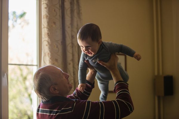 An older man joyfully lifts a smiling baby indoors near a window.