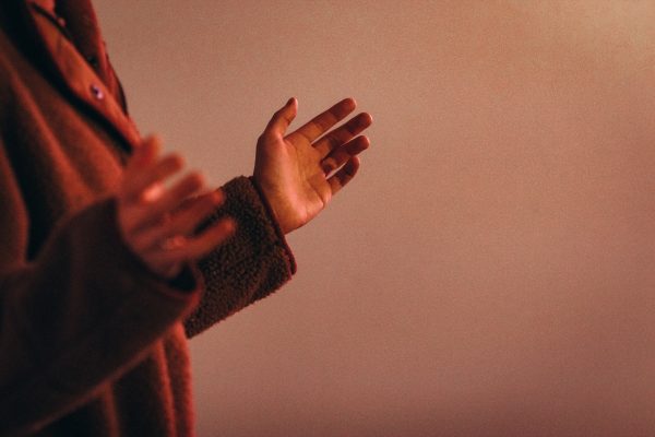 Person in a warm brown sweater gesturing with hands against a plain, muted background.