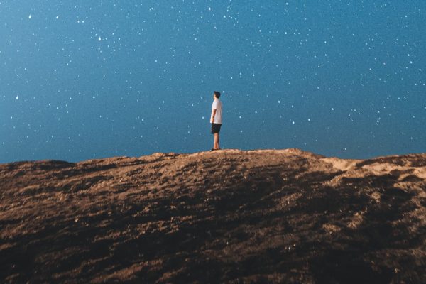 person standing small on rocky land beneath blue sky full of stars