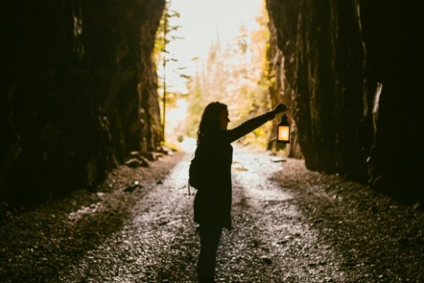 Silhouette of a person holding a lantern inside a tunnel with light at the end.