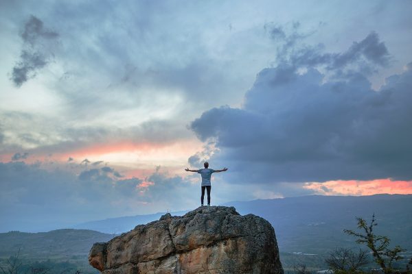 Person standing on a rock with arms outstretched, overlooking a scenic sunset with dramatic clouds.