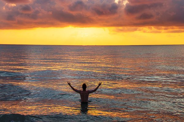 person standing in ocean water with arms raised to yellow-pink sky, sun rising or setting