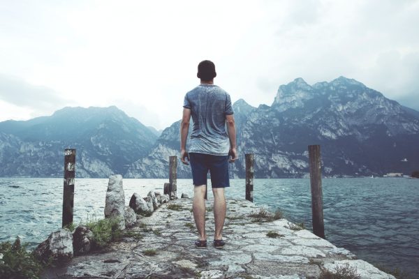 A man stands on a rocky pier, facing a lake with large mountains in the background under a cloudy sky.