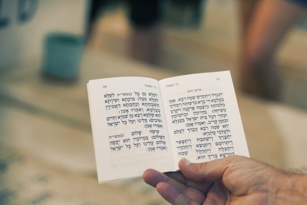 A hand holding an open book with Hebrew text, blurred background.