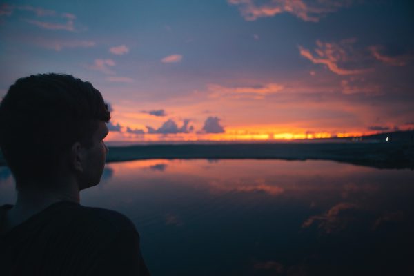 Silhouette of a person gazing at a vibrant sunset over a calm lake with colorful clouds in the sky.