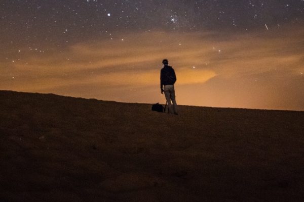 Person with a tripod on a desert at night, gazing at the starry sky with scattered clouds.