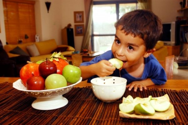 Child eating green apple by a table with a fruit bowl in a cozy living room.