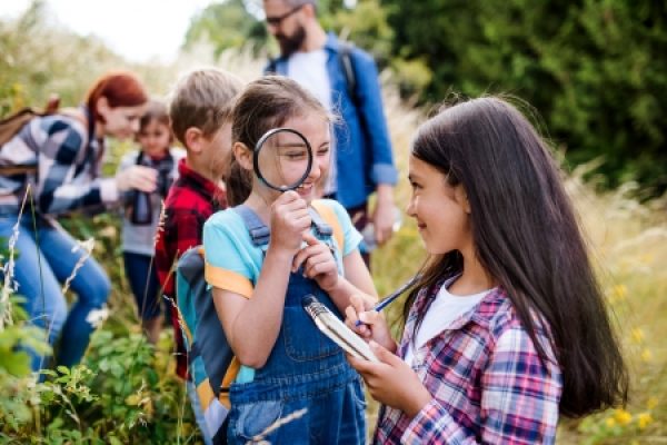 Children explore nature with a magnifying glass and notebook, guided by adults in a grassy outdoor setting.