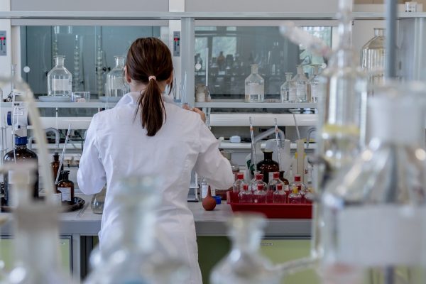 A person in a lab coat works at a laboratory bench with various bottles and equipment.