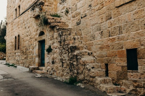 Stone building with weathered steps and a blue door on a narrow street. Vegetation grows in cracks.