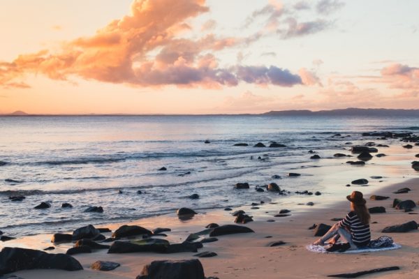 Person sitting on a beach at sunset, surrounded by rocks, with a colorful sky and calm ocean in the background.