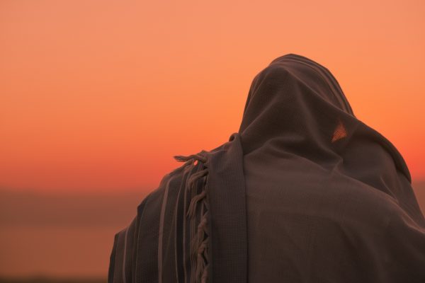 Person draped in a cloth facing a serene orange sunset sky.