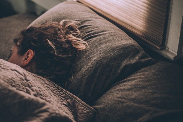 Person sleeping peacefully under a brown blanket beside a window with blinds.